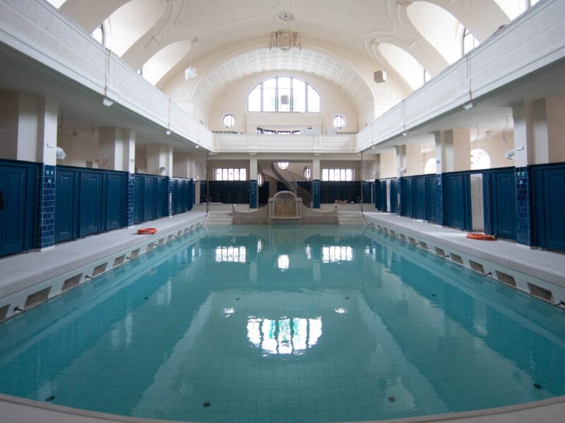 Vue sur le Petit Bassin des Bains Municipaux de Strasbourg. Une piscine est dans une salle voutée baignée de lumière naturelle par des oeil de boeuf latéraux et un vitrail. Une coursive court à l'étage. Le plafond vouté et les murs sont blancs, les cabines qui entourent la piscine sont bleu roi. Au fond de l'image, on voit une fontaine entre les deux escaliers qui permettent d'entrer dans la piscine.