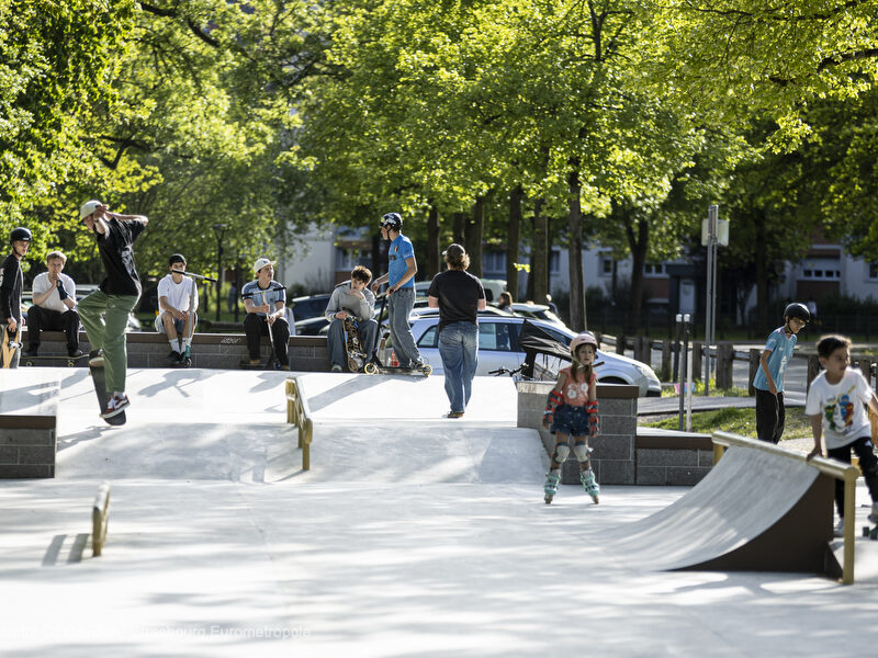 Plusieurs riders assis sur la partie haute observent le skatepark : on voit des modules en escalier, des pentes en béton lisse, une rampe de slide.