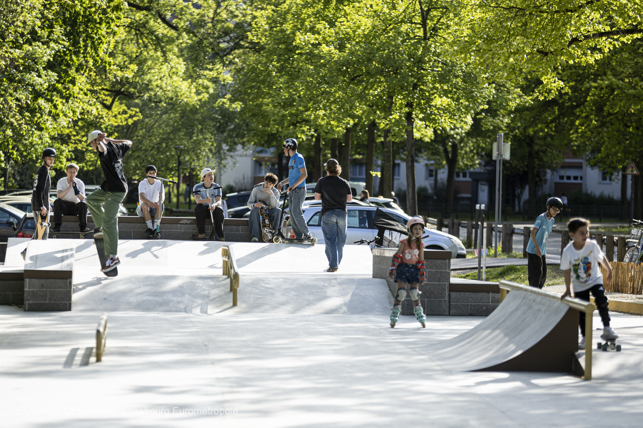 Plusieurs riders assis sur la partie haute observent le skatepark : on voit des modules en escalier, des pentes en béton lisse, une rampe de slide.