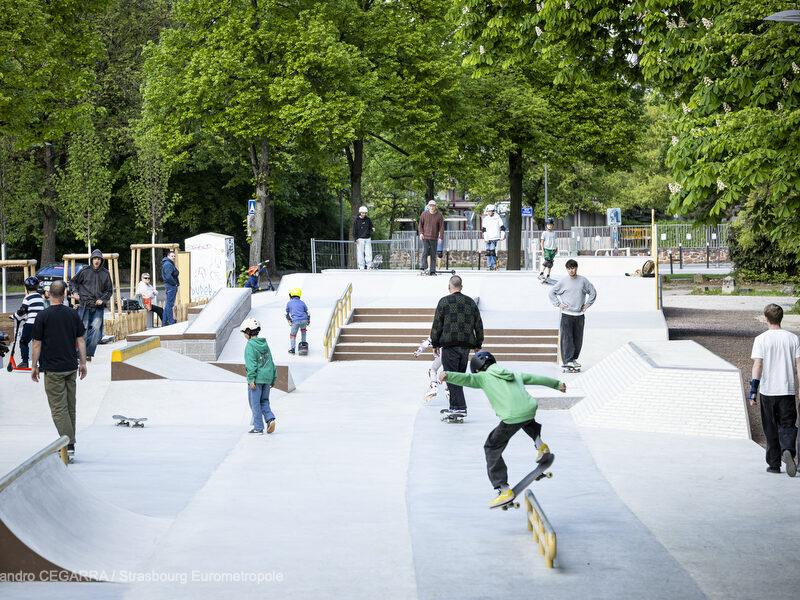 Plusieurs riders en train de rouler sur le skatepark : on voit des modules en escalier, des pentes en béton lisse, une rampe de slide sur lequel un jeune skater en pull vert slide.