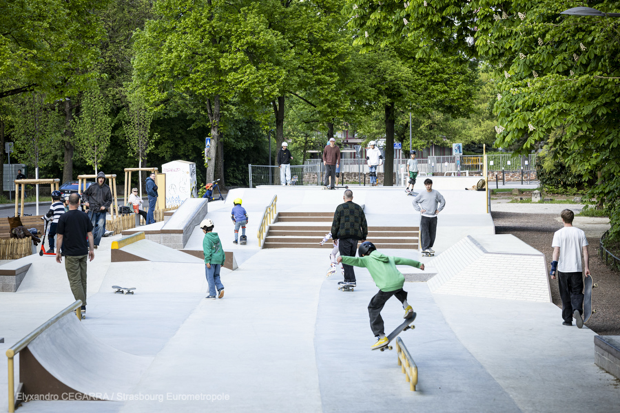 Plusieurs riders en train de rouler sur le skatepark : on voit des modules en escalier, des pentes en béton lisse, une rampe de slide sur lequel un jeune skater en pull vert slide.