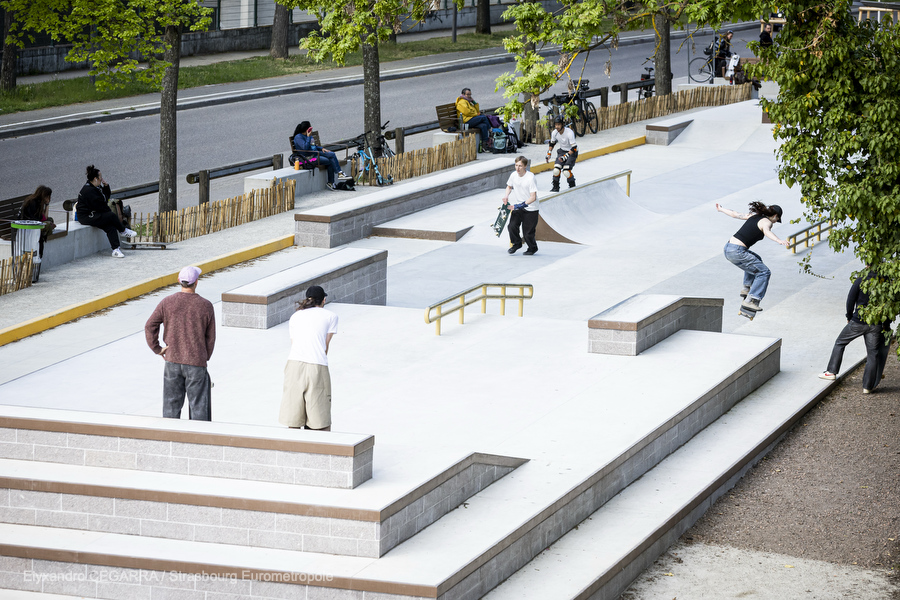 Vision d'ensemble du skatepark : des modules en escalier, des pentes en béton lisse, une rampe de slide. On voit la rue de Boston qui longe l'équipement et les assises installées le long