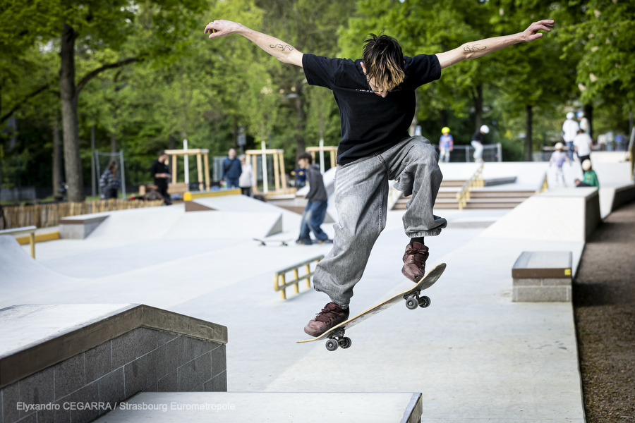 Vision d'ensemble du skatepark : des modules en escalier, des pentes en béton lisse, une rampe de slide. Un skater effectue un saut au premier plan