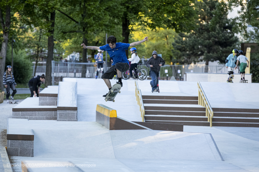 Vision d'ensemble du skatepark : des modules en escalier, des pentes en béton lisse, une rampe de slide. Un skater effectue un saut au premier plan